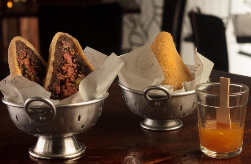 Foto de comida de boteco. Em cima de um balcão de madeira há duas pequenas travessas de alumínio, com uma coxinha em cada. A da esquerda, a coxinha está aberta e dá para ver o recheio de feijoada. A da direita está inteira. À frente delas há um pequeno copo com um molho e uma espátula de madeira. 