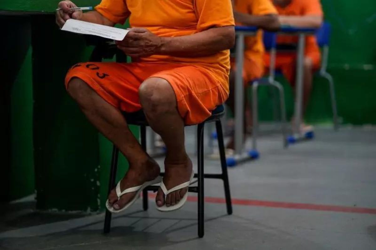 Imagem de detentos, vestindo uniformes laranjas, sentados em uma mesa de sala de aula, para contextualizar com o fato de que muitas salas da nova unidade serão focadas em estudos e trabalhos para a ressocialização dos prisioneiros 