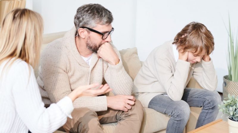 Homem grisalho e mulher loira discutindo no sofá e criança tampando os ouvidos