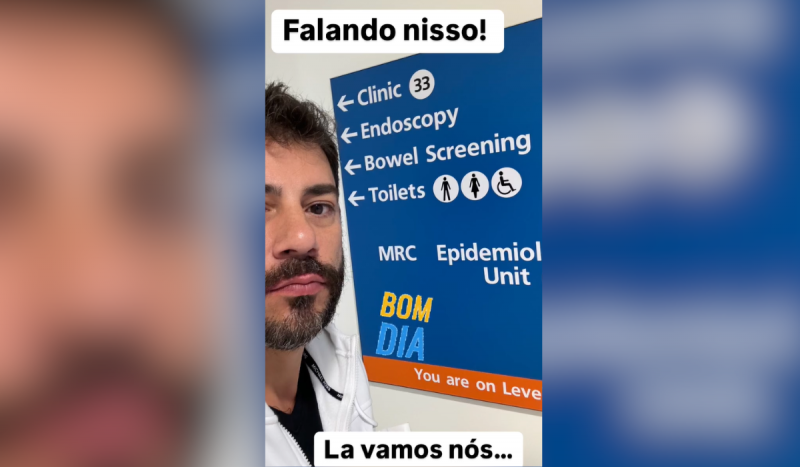 Captura de tela mostra homem branco, de barba e bigode, usando camiseta preta e moletom branco por cima. Ele está ao lado de placa azul com indicações de salas em hospital na Inglaterra, as palavras estão em inglês