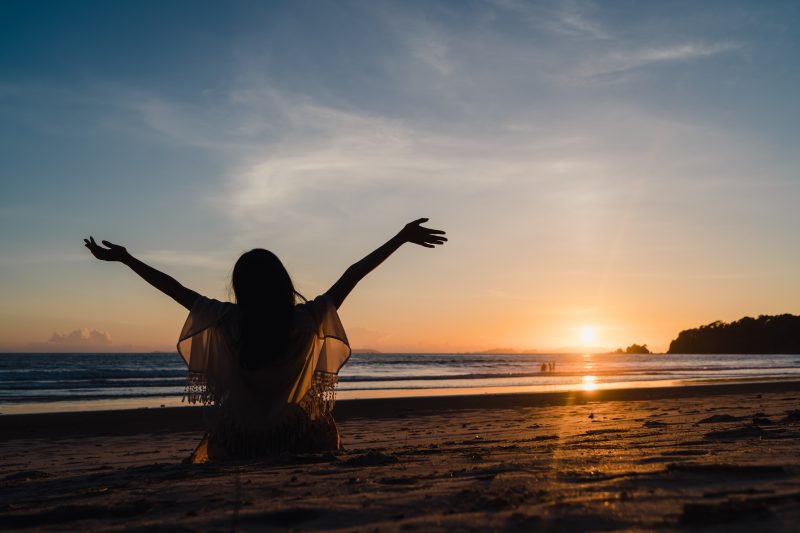 Mulher na praia com os braços abertos para o mar e o sol 