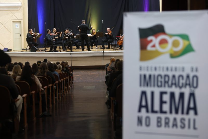 Orquestra Prelúdio no palco do teatro ao fundo, na frente um banner com logo do projeto Bicentenário da Imigração Alemã