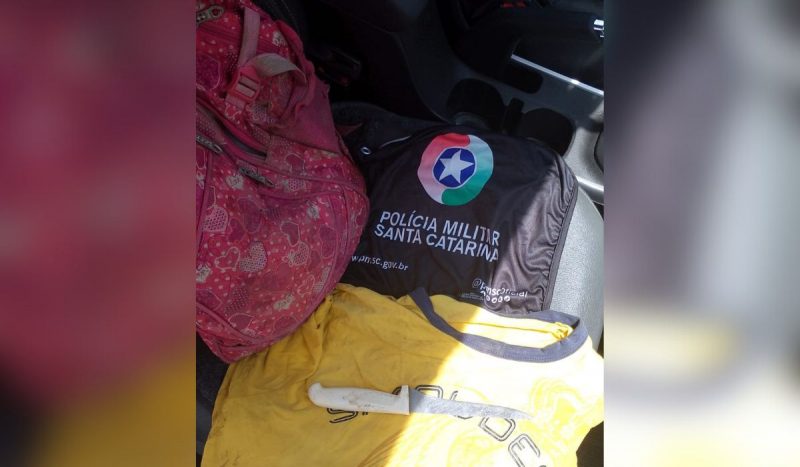 Faca em cima de blusa amarela suja, ao lado de bandeira com insígnia da PMSC para ilustrar matéria de homem esfaqueado em confusão em bar