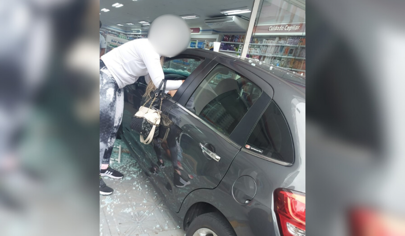 Carro cinza com parte dianteira dentro de farmácia após colisão. Vidros estilhaçados estão espalhados no chão e mulher branca de cabelos loiros e longos está debruçada na janela do motorista. Acidente ocorreu em São José