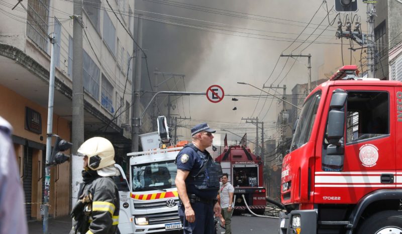 Foto mostra policial andando na rua ao lado de viatura dos bombeiros. Ao fundo, fumaça intensa de incêndio no Brás pode ser vista subindo