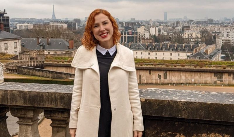 Influenciadora brasileira que morreu atropelada na França - mulher branca, ruiva, de cabelos curtos e cacheados, posa em pé sorrindo enquanto usa uma blusa preta com gola branca e casaco sobretudo na cor bege. Ela está em frente à paisagem na França