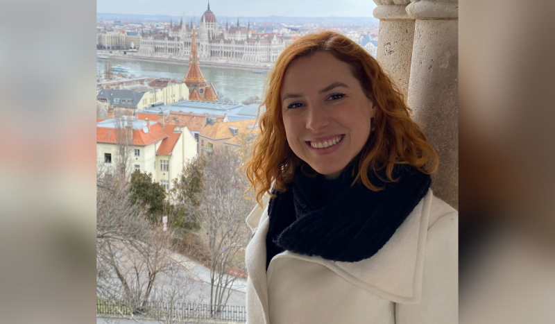 Mulher branca, ruiva, de cabelos curtos e ondulados, sorri com paisagem na França de fundo. Ela usa cachecol preto com casaco bege 