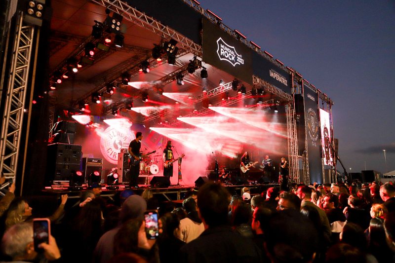 A foto noturna mostra um palco de forma lateral. Em cima dele há cinco músicos e vários focos de luzes vermelhas. Em cima do palco está escrito: Mundial Rock 2023. À frente dele, há dezenas de pessoas, algumas tirando fotos com o celular. 