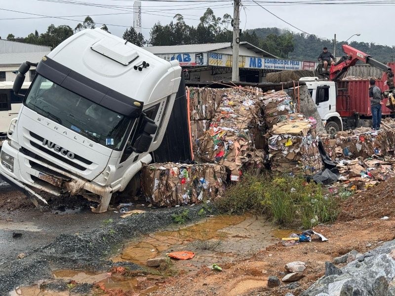 A imagem mostra um caminhão tombado com materiais recicláveis em Blumenau.