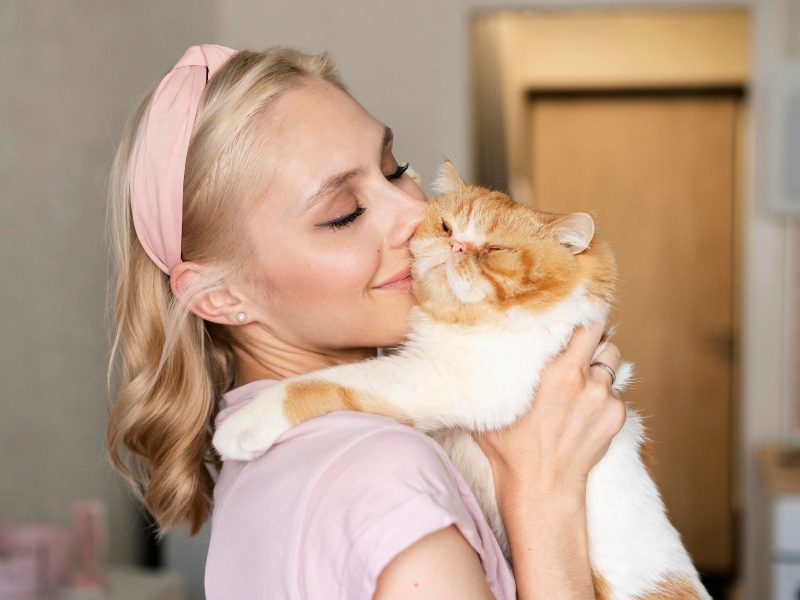 A imagem mostra uma mulher loira com uma faixa rosa no cabelo segurando um gato branco e laranja no colo.