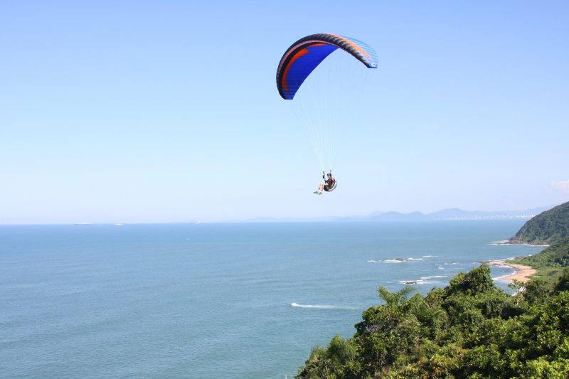 Pessoa sobrevoando o mar em um parapente. No canto direito da foto aparece vegetação e ao fundo um pedaço de areia. 