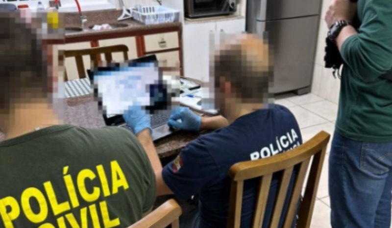 Operação prende seis pessoas suspeitas de armazenar pornografia infantil em SC, foto mostra policiais sentados em uma mesa na cozinha enquanto um deles mexe de luvas em um notebook