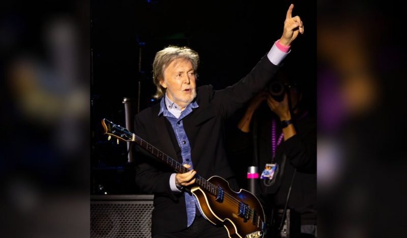 Paul McCartney levantando o braço esquerdo em direção ao público em show - homem branco, de cabelos lisos e grisalhos, usa camisa azul com jaqueta preta por cima enquanto toca baixo