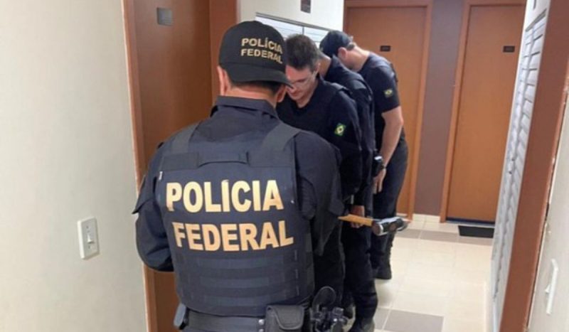 Quadrilha suspeita de enviar drogas para o Aeroporto de Florianópolis é desarticulada no DF, foto mostra policiais fardados em frente a uma porta fechada. Um deles carrega uma marreta