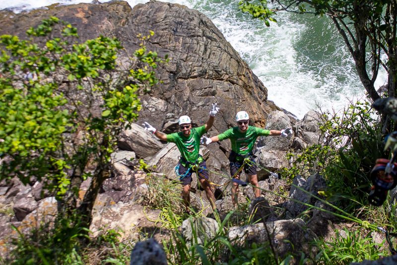 Dois homens estão amarrados por uma corda e descem um costão de rochas. Eles sorriem para a foto e estão com os braços erguidos. Ao fundo, aparece um pedaço do mar. 