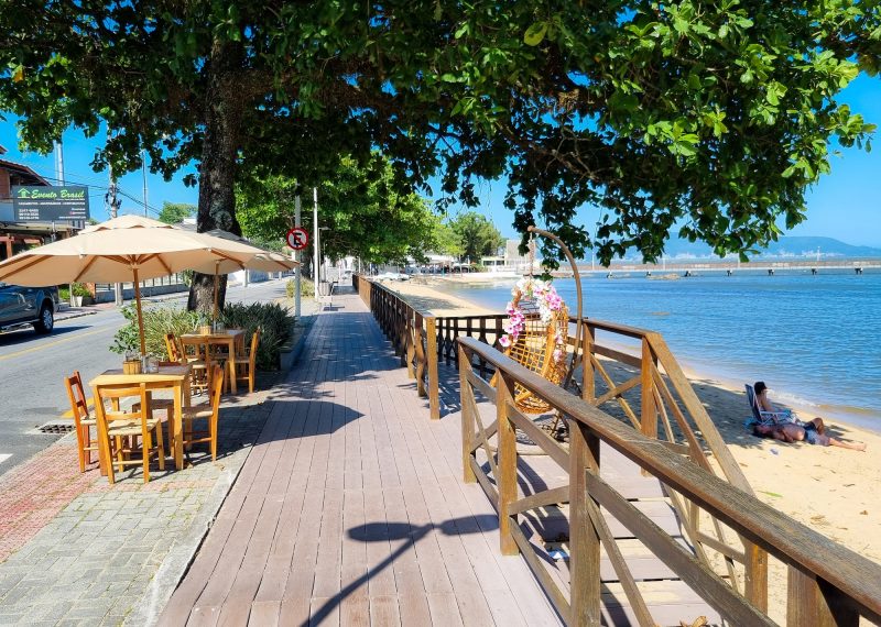 A foto mostra uma passarela de madeira ao lado de uma calçada. Na calçada, há duas mesas de madeira com cadeiras e uma árvore bem grande. Do lado direito da foto aparece uma faixa de areia e as águas do Balneário de Guararema. Ao fundo, aparece um trapiche de concreto e mais árvores ao redor da areia. 