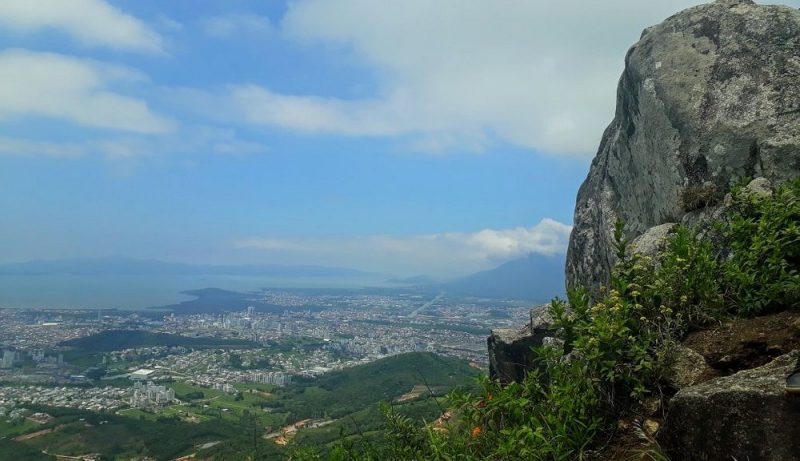 Foto aérea da cidade e ao fundo o mar. No canto direito da foto há uma grande pedra. O céu está azul. 