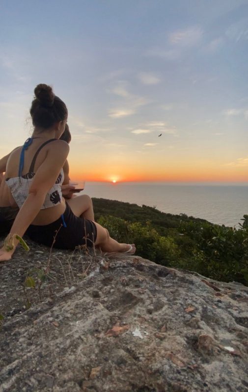 Casal observa o sol. Eles estão sentados em uma pedra, no Farol da Lagoa, uma das trilhas de Florianópolis