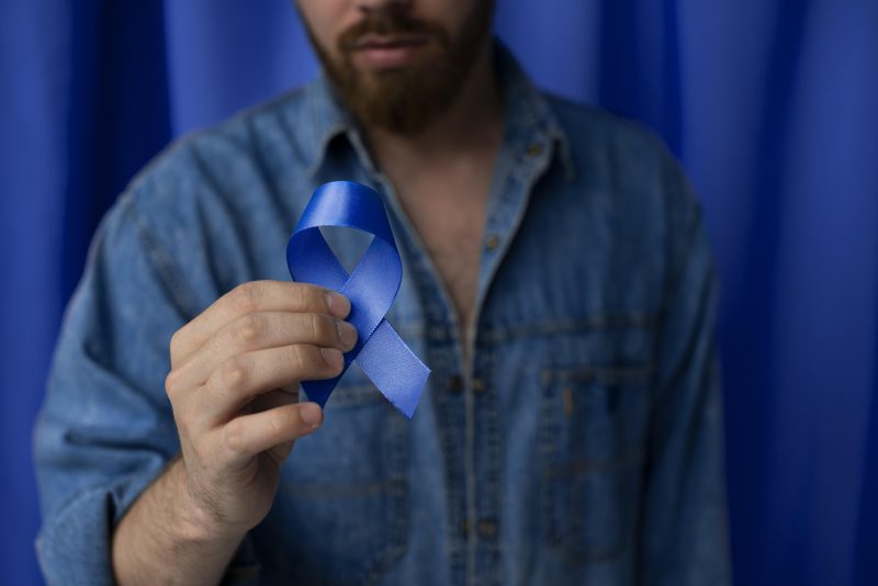 Homem desfocado ao fundo, usando camisa jeans azul e segurando um laço azul 