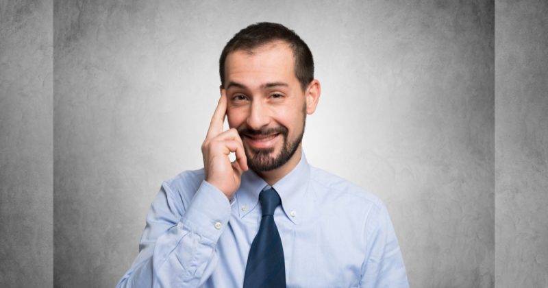 Homem sorridente de blusa social e gravata com dedo na testa