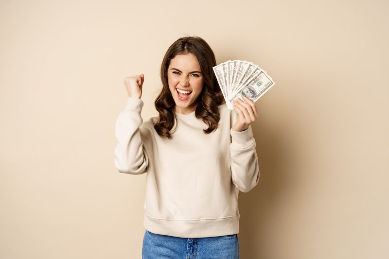 mulher com um leque de dólares na mão esquerda fazendo sinal de comemoração com a mão direta e sorrindo remetendo aos que podem ficar ricos na loteria