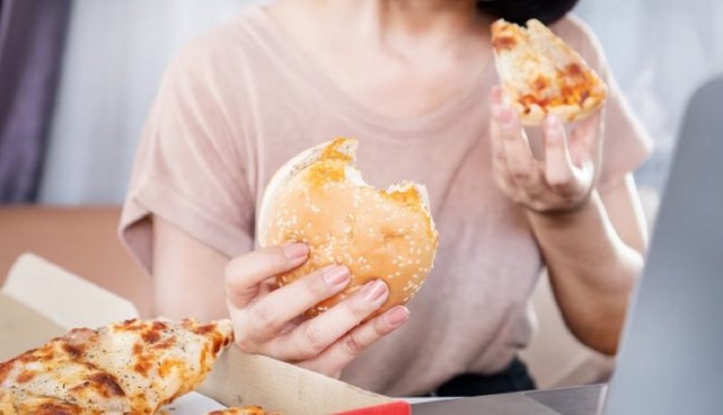 na foto aparece uma pessoa com hamburguer e pizza na mão e que pode ter problemas por comer rápido demais