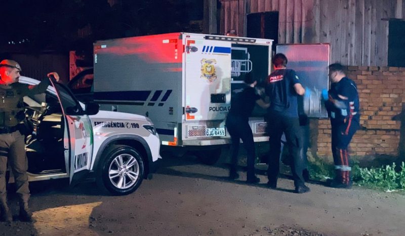 foto mostra três agentes colocando corpo de homem morto a tiros em veículo da polícia científica, enquanto policial militar observa de viatura