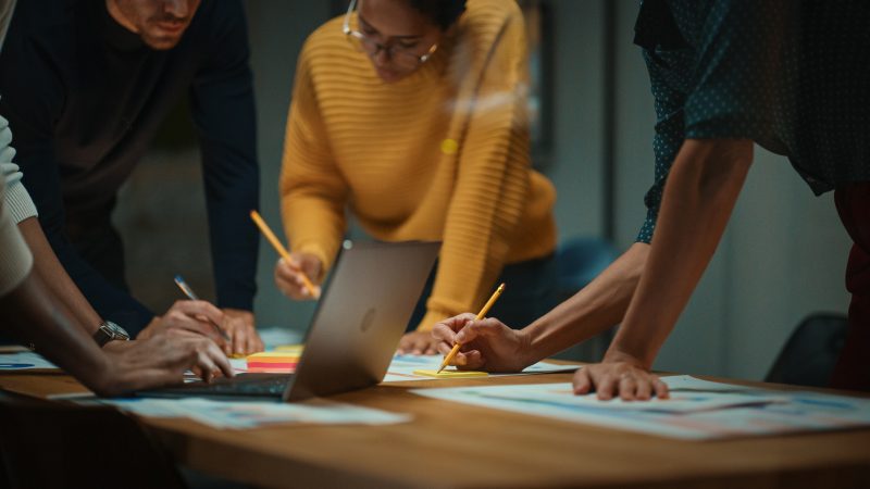 Colegas se inclinam em uma mesa de conferência, olham para o computador portátil e fazem anotações de um planejamento de mídia com lápis e notebooks.