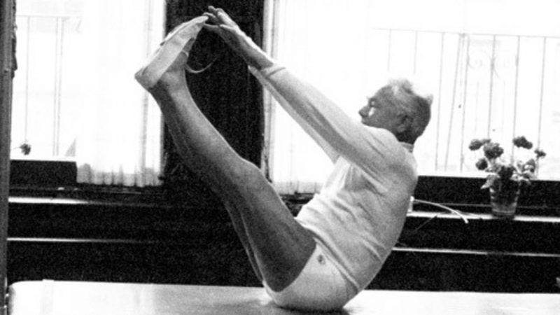 Imagem antiga de Joseph Pilates, um homem branco se alongando, segurando as pontas dos pés enquanto estando sentado