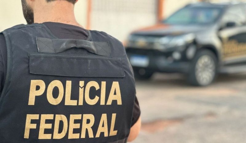 Policial federal de costas com o símbolo da corporação nas costas e carro ao fundo 