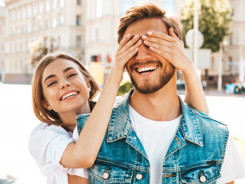 casal sorrindo: mulher atrás do homem com as mãos tapando os olhos dele e ele em primeiro plano com uma camiseta branca e um colete jeans