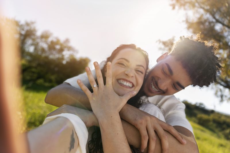 casal sorrindo sentado na grama ele está atrás dela e a abraça pelo pescoço enquanto sorri e olha para ela; já a mulher mostra a mão com uma aliança enquanto sorri representando os signos que combinam com escorpião 