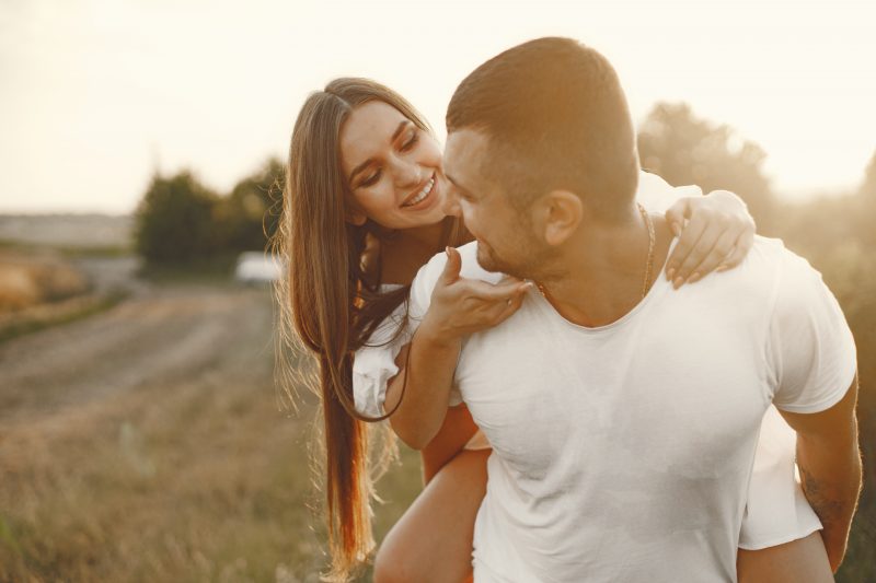 casal em um campo aberto, ambos de roupa branca e o homem está carregando a mulher nas costas; ambos estão se olhando e sorrindo 