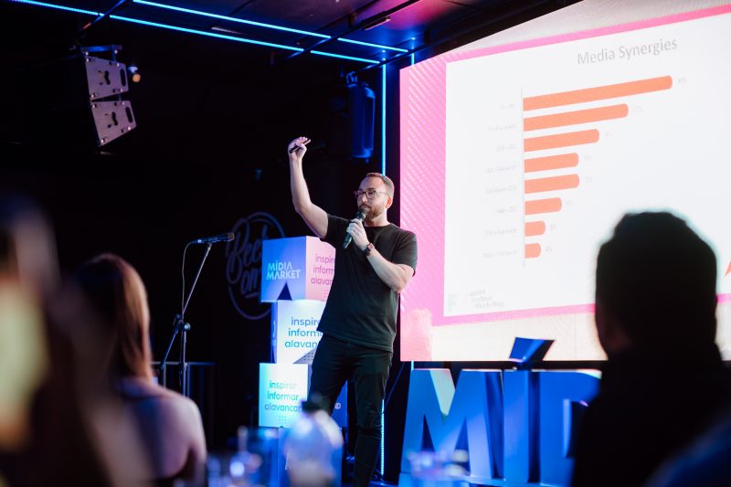 Homem vestido todo de preto está sob um palco e fala para o público. Ele segura um microfone com uma das mãos e com a outra ergue o passador de slides.