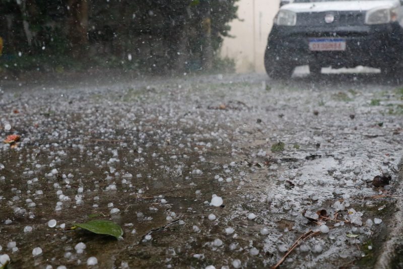 Imagem de chuva de granizo caindo no chão com um carro ao fundo