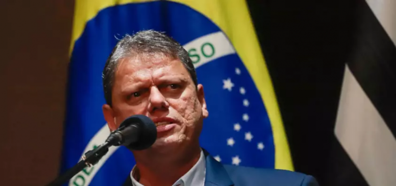 Tarcísio de Freitas falando no microfone com bandeira do Brasil ao fundo ao lado de bandeira do estado de São Paulo