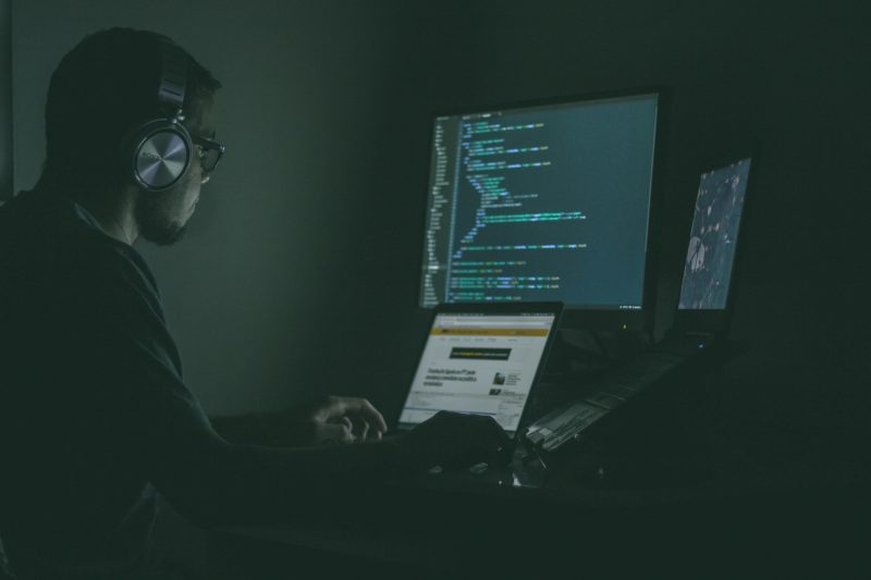 Homem com fones de ouvindo trabalhando como programador em frente a três computadores 