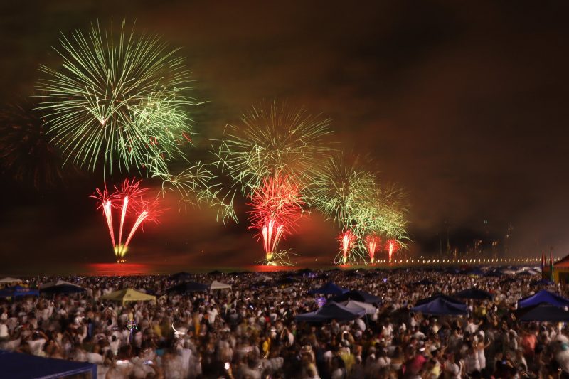 Foto da queima de fogos de artifício na Praia Central de Balneário Camboriú no Ano Novo. A areia da praia está completamente cheia com milhões de pessoas. Em alto mar, os fogos têm cores de verde e vermelho, em formato circular. 