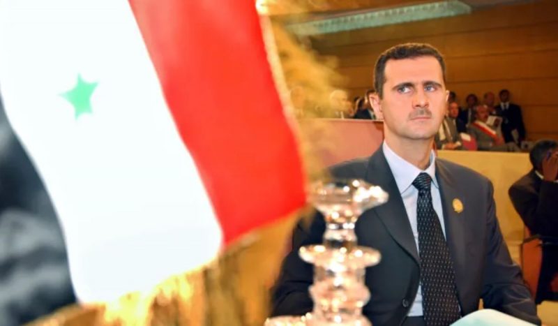 Foto mostra Bashar al-Assad sério com bandeira da Síria na frente