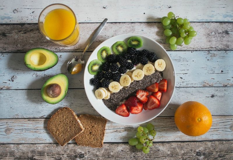 Imagem de uma tigela de com frutas, como kiwi, banana, morango, amora, acompanhada de suco de laranja e outras frutas na mesa 