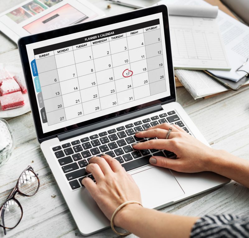 Computador em cima de uma mesa aberto num calendário com as mãos de uma pessoa no teclado e diversos papéis sobre a mesa