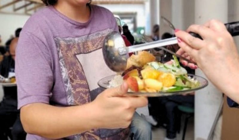 Cozinha solidária costumava distribuir, gratuitamente, 400 refeições vegetarianas por semana, foto mostra pessoa de camiseta roxa segurando prato cheio de comida enquanto pessoa a serve