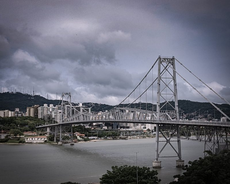 Foto da ponte Hercílio Luz, toda em estrutura metálica, passando por cima das águas da baía Norte e Sul de Florianópolis. Ao fundo, prédio altos e morros verdes. O céu está cinza e carregando de nuvens. Imagem ilustrativo do que fazer em Florianópolis quando chove. 