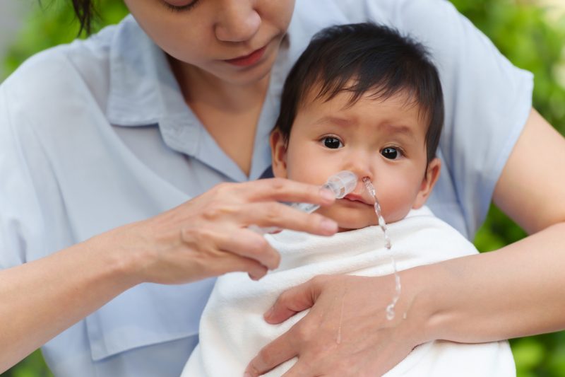 Bebê asiático no colo de uma mulher que faz lavagem nasal na criança com auxilio de uma seringa 