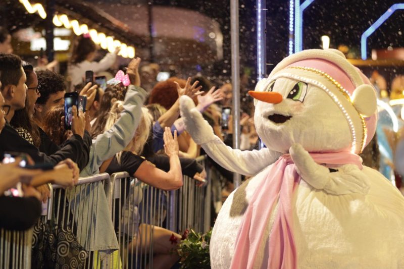 Pessoa fantasiada de boneco de neve acena para plateia que assiste ao desfile de rua do Natal em Blumenau. 