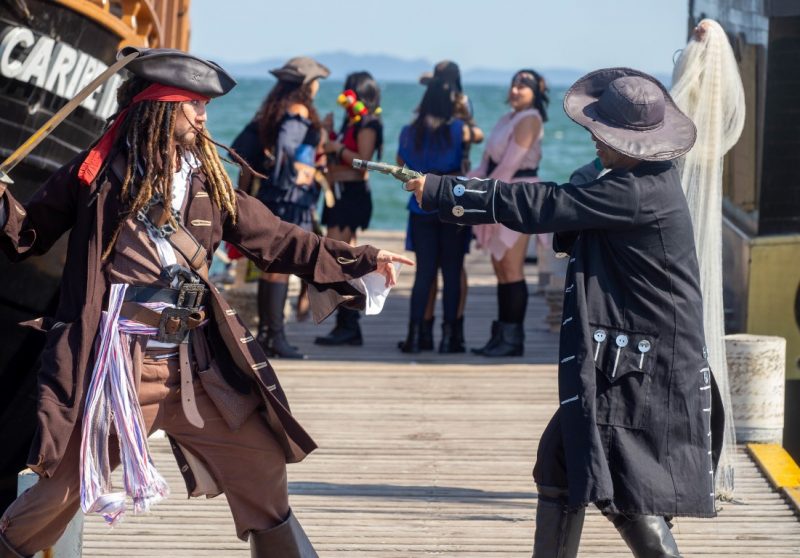 Foto de dois homens jovens vestidos de pirata sobre uma passarela de madeira. Um deles tem uma espada na mão e o outro um arma. Eles estão de frente um para o outro, de forma lateral para a foto, e apontam as armas um para o outro. Ao fundo estão outros personagens do passeio temático e o mar. No canto esquerdo da foto é possível ver uma parte do barco pirata. 