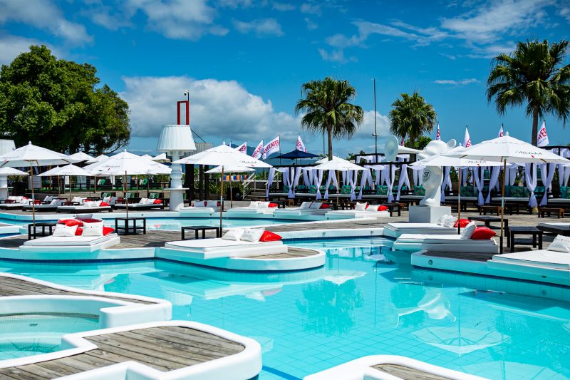 Foto interna do beach club P12. Aparecem vários decks onde estão colchões, com mesinhas e guarda-sóis sobre uma grande área de piscina. Ao fundo, aparecem alguns coqueiros e gazebos. O céu está azul. 