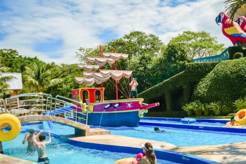 Foto de uma área do Água Show Park, em Ingleses - Florianópolis. Aprecem duas piscinas rasas com algumas crianças. Uma pequena ponte une as duas piscinas. Em uma delas há um pequeno barco a velas. Ao redor das piscinas, há algumas árvores e duas grandes esculturas de araras. O céu está bem azul e o dia ensolarado. 