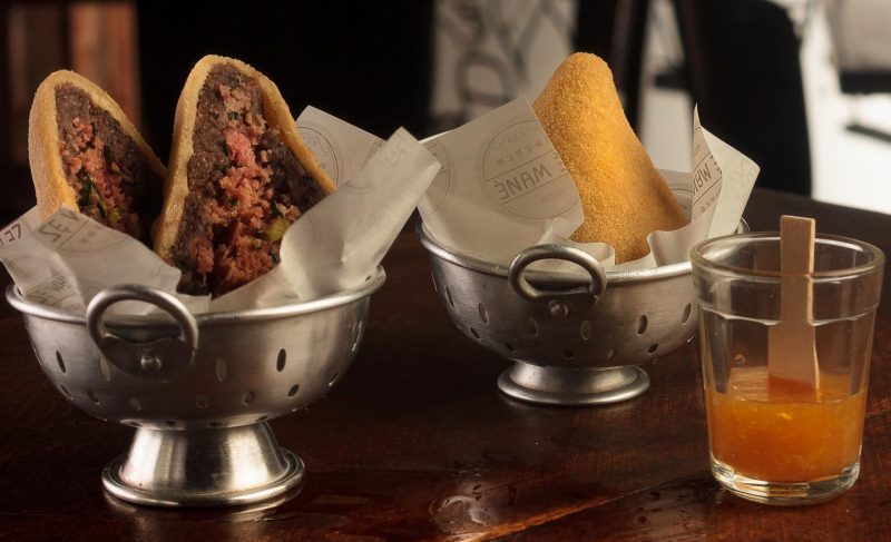 Foto de uma coxinha cortada ao meio, com o recheio de feijoada à mostra. Ao lado uma coxinha inteira. As duas estão em pequenos escorredores de alumínio onde são servidas, em cima de um balcão de madeira do Boteco Zé Mané. 