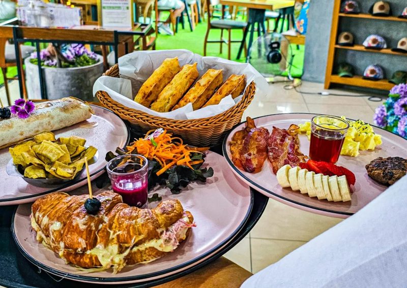 Foto de vários pratos do Bulegreen Café & Brunch, como croissant, bacon, ovos mexidos e frutas. Eles estão em bandejas em cima de um balcão de madeira. 